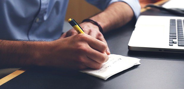 A photograph of a man writing on a piece of paper next to an open laptop. 