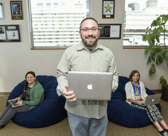Belasco_Team A photograph of Mike Belasco standing and holding an open laptop. Behind him, Delia Valdez and Gina Kuzmick sit on bean bags with open laptops on their lap.