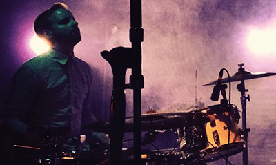 A photograph of Neil Robertson playing drums on concert stage.