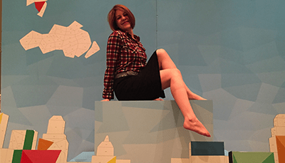 A photograph of Sara Downey Robinson sitting on top of a block behind a constructed city scape. 