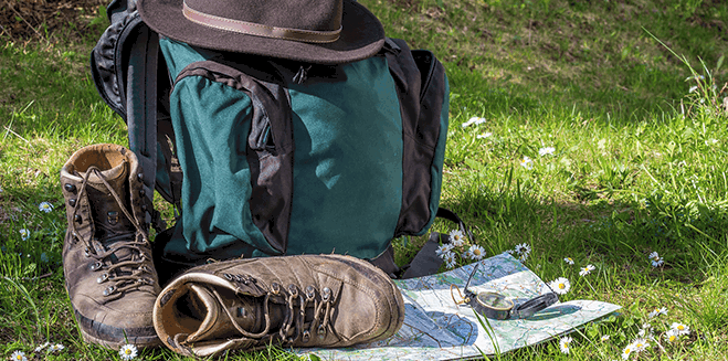 A photograph outdoors of a backpack with a hat on top of it. Next to it are a pair of hiking boots, a map and a compass. 