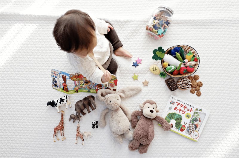 A photograph from above of a young child sitting on a rug surrounded by toys. 