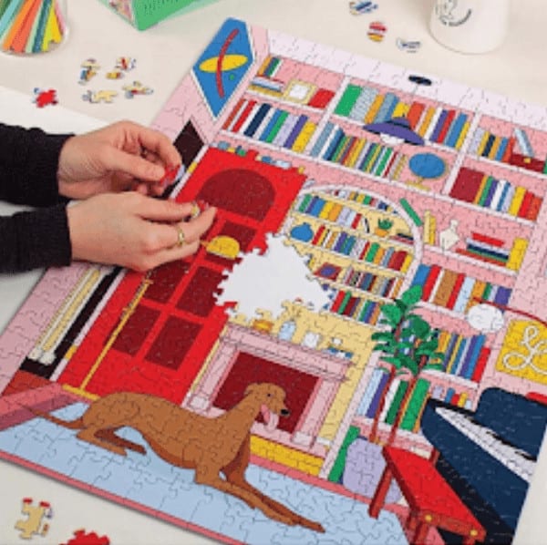 A photograph of a person assembling a brightly colored puzzle of a dog lying in a room with books and a piano. 