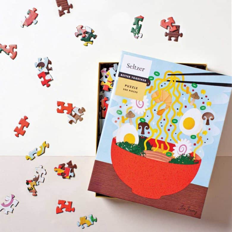 Ramen-Puzzle A photograph of a slightly opened, brightly colored Seltzer puzzle box with an image of ramen on the cover. Puzzle pieces surround the box.