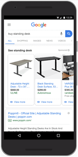 Google-Shopping-ads-example-1 A photograph of a smartphone displaying Google shopping ads for the search text "buy standing desk." Ads are at the top of the search results. Each ad contains an image of the product and no text description.