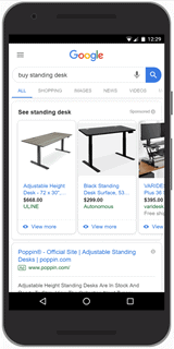 A photograph of a smartphone displaying Google shopping ads for the search text "buy standing desk." Ads are at the top of the search results. Each ad contains an image of the product and no text description. 