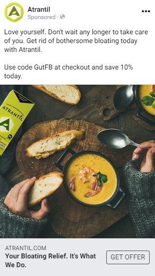 An Atrantil Facebook ad.  A short text advertises Atrantil as getting rid of bloat and offers a discount code. Below the text is a photograph of a person wearing a sweater and eating a bowl of yellow soup containing shrimp and toast served on a dark wood serving platter. A package of Atrantil is nearby. 