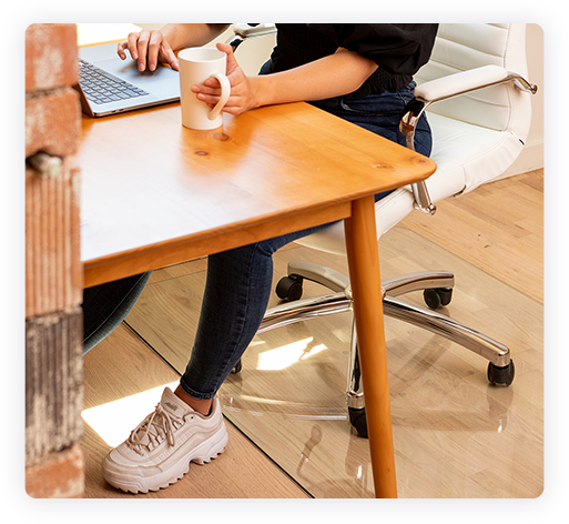 A photograph of a person sitting in an office chair at a desk working on a laptop and holding a coffee. A glass mat is under the chair. 